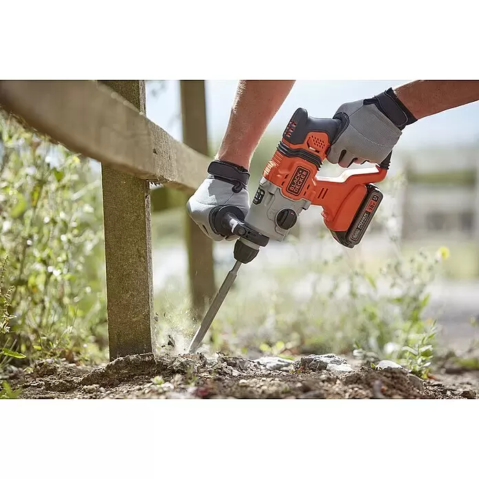 Black+Decker Akku-Kombihammer BCD900E2K-QW - 18 V, 2 Akkus, 1,2 J - Eine Person mit Handschuhen benutzt einen orangefarbenen und grauen Black+Decker Akku-Bohrhammer mit Meißel, um den Boden neben einem Holzpfosten aufzubrechen.