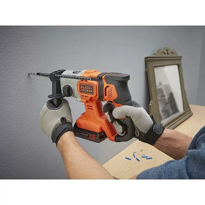Black+Decker Martillo combinado de batería BCD900E2K - 18 V, 2 baterías, 1,2 J - Persona taladrando una pared gris con un taladro inalámbrico BLACK+DECKER naranja y negro, usando guantes de trabajo.