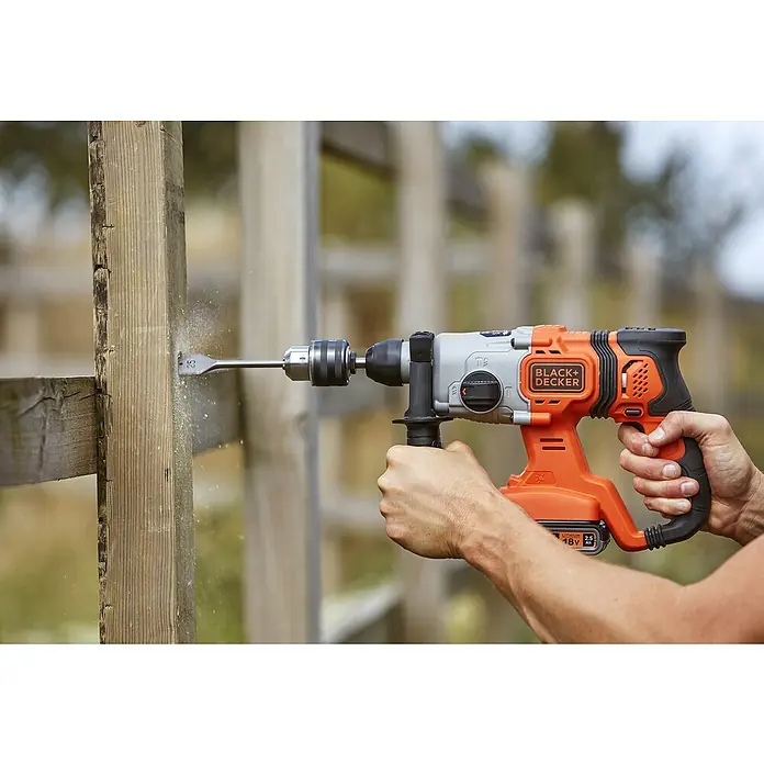 Black+Decker Akku-Kombihammer BCD900E2K-QW - 18 V, 2 Akkus, 1,2 J - Ein Black+Decker Akku-Bohrhammer wird benutzt, um ein Loch in einen Holzzaun zu bohren. Das Werkzeug ist orange und grau.