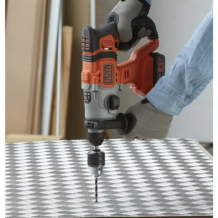 Black+Decker Akku-Kombihammer BCD900E2K-QW - 18 V, 2 Akkus, 1,2 J - Akku-Bohrmaschine von Black+Decker in Aktion, die ein Loch in ein Metallstück bohrt, gehalten von Händen mit Arbeitshandschuhen für sicheren Halt.