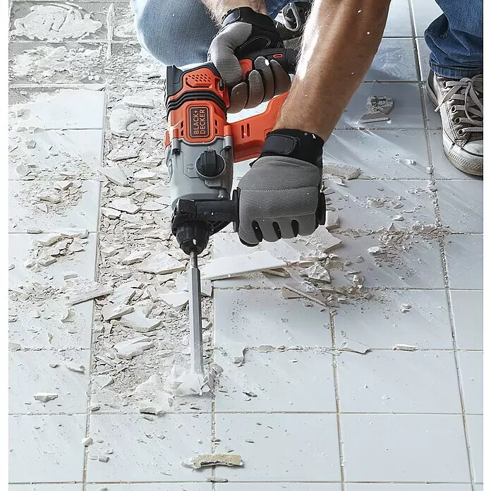 Black+Decker Akku-Kombihammer BCD900E2K-QW - 18 V, 2 Akkus, 1,2 J - Person verwendet einen orange-schwarzen BLACK+DECKER Bohrhammer mit Meißel zum Aufbrechen weißer Bodenfliesen, trägt graue Arbeitshandschuhe.