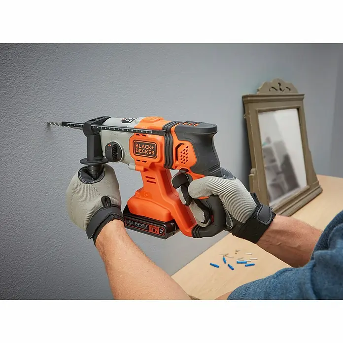 Black+Decker Martillo combinado de batería BCD900B - 18 V, Sin batería, 1,2 J - Un manitas perfora un agujero en una pared gris con un taladro percutor Black+Decker. El taladro es naranja y negro.