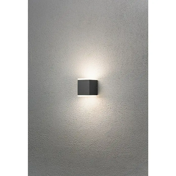 Konstsmide Außenwandleuchte - 12 W, 9 x 6 x 15 cm, Schwarz, IP54 - Diese Aufnahme zeigt eine moderne, graue Wandleuchte, die ein warmes Licht an eine strukturierte Wand wirft. Das Design ist minimalistisch und elegant.