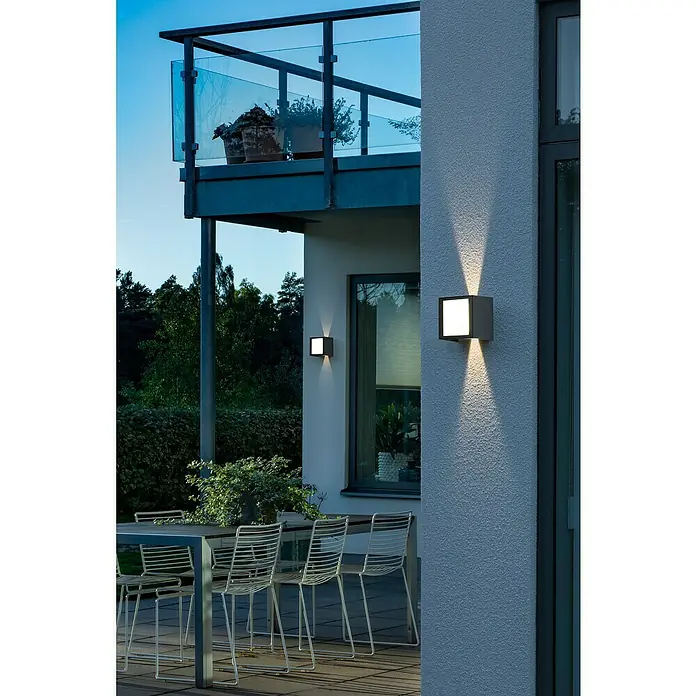 Konstsmide LED-Außenwandleuchte - 8 W, 8 x 16,5 x 11,5 cm, Weiß, IP54 - Eine moderne Außenwandleuchte spendet warmes Licht und akzentuiert die Fassade eines modernen Hauses mit Balkon und Gartenmöbeln.
