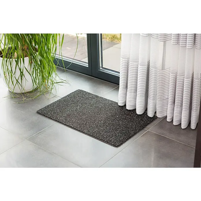 Astra Fußmatte Rib Line Sprint - 50 x 80 cm, Anthrazit, 100 % Polypropylen - Graue Fußmatte vor einer Tür, neben einer Zimmerpflanze und einem weißen Vorhang.
