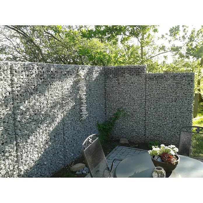 Gabiona Gabione - 107 x 17 x 180 cm, Metall, Maschenbreite: 50 mm, Maschenhöhe: 50 mm - Eine Gabionenwand umgibt eine Terrasse mit Tisch und Stühlen, geschmückt mit Pflanzen. Die Gabionen sind mit Steinen gefüllt und bieten Sichtschutz.