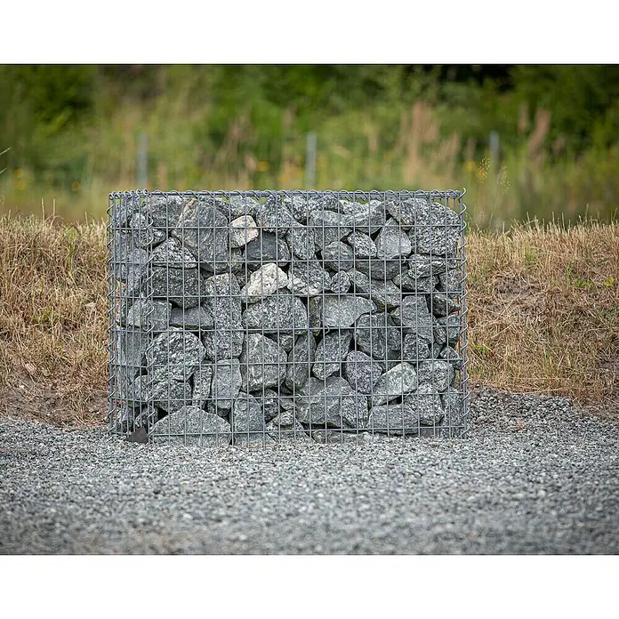 Gabiona Gabione - 100 x 30 x 80 cm, Metall, Maschenbreite: 50 mm, Maschenhöhe: 100 mm - Eine Gabione, ein Drahtkorb gefüllt mit grauen Steinen, steht auf einem Kiesweg vor einer grünen Graslandschaft.