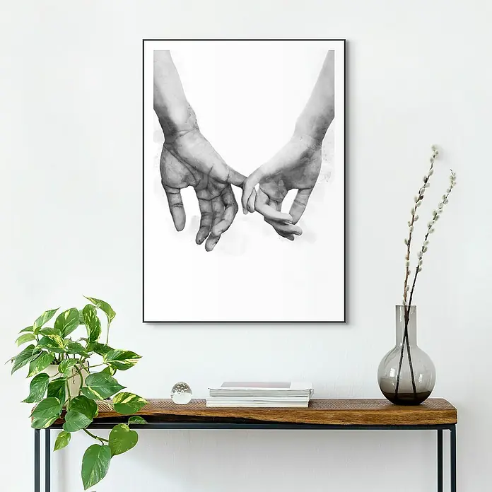 Reinders Bild Slim Frame BlackPinkies, B x H: 50 x 70 cm - Das Schwarz-Weiß-Foto zeigt ein gerahmtes Bild von zwei Händen, die sich zärtlich berühren, und steht auf einem stilvollen Holztisch neben Dekorationen.