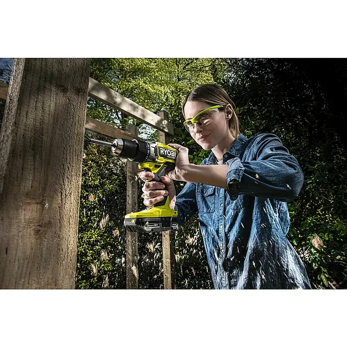 Ryobi ONE+HP Taladro atornillador de batería RDD18X - 18 V, Sin batería, 95 Nm - Una mujer está perforando un agujero en una tabla de madera con un taladro inalámbrico Ryobi. Hay partículas de polvo volando alrededor.