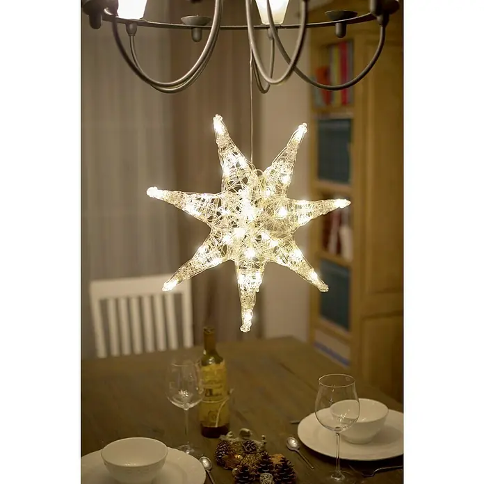 Konstsmide LED-Stern - 45 cm, Farblos, Lichtfarbe: Warmweiß, IP20, Netzbetrieben - Ein sternförmiges Dekorationsobjekt mit Lichtern hängt über einem Tisch mit Gedeck, Wein und Tannenzapfen.