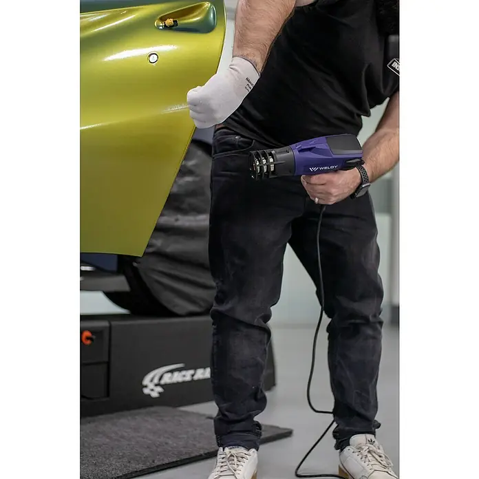 Weldy Heißluftgebläse HG 330-B Car-Wrap-Kit - 1.600 W, Temperaturbereich: 350 °C - 550 °C - Nahaufnahme eines WELDY-Werkzeugs in Aktion, das bei professionellen Reparaturarbeiten an Autos verwendet wird.