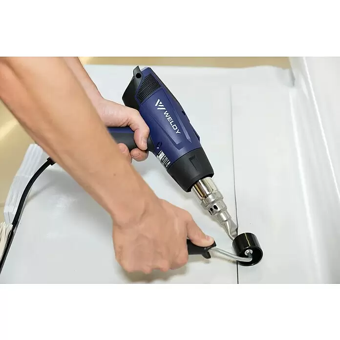 Weldy Heißluftgebläse HG 530-A - 2.300 W, Temperaturbereich: 80 °C - 650 °C - Hände nutzen eine blau-schwarze WELDY Heißluftpistole mit Andrückrolle zum Schweißen/Abdichten von hellem Material.