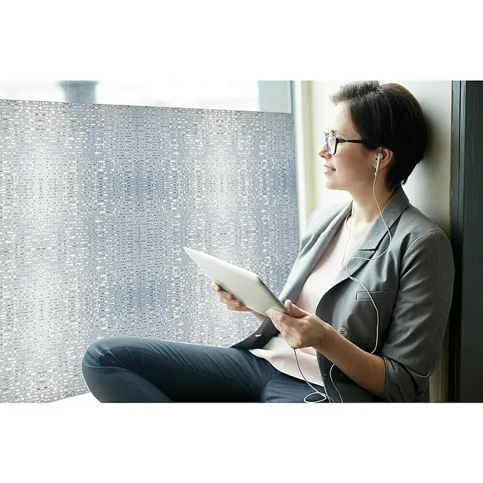 Lámina para ventana al corte Viena - Ancho: 92 cm, Transparente - Una mujer con gafas y auriculares sentada junto a la ventana sosteniendo una tableta, con una película decorativa en la ventana.