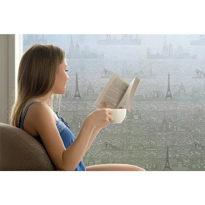 Lámina para ventana Viajes - 92 cm x 1,5 m, Transparente - Una joven se relaja con una taza de café y un libro frente a un vinilo para ventanas con el horizonte de París.
