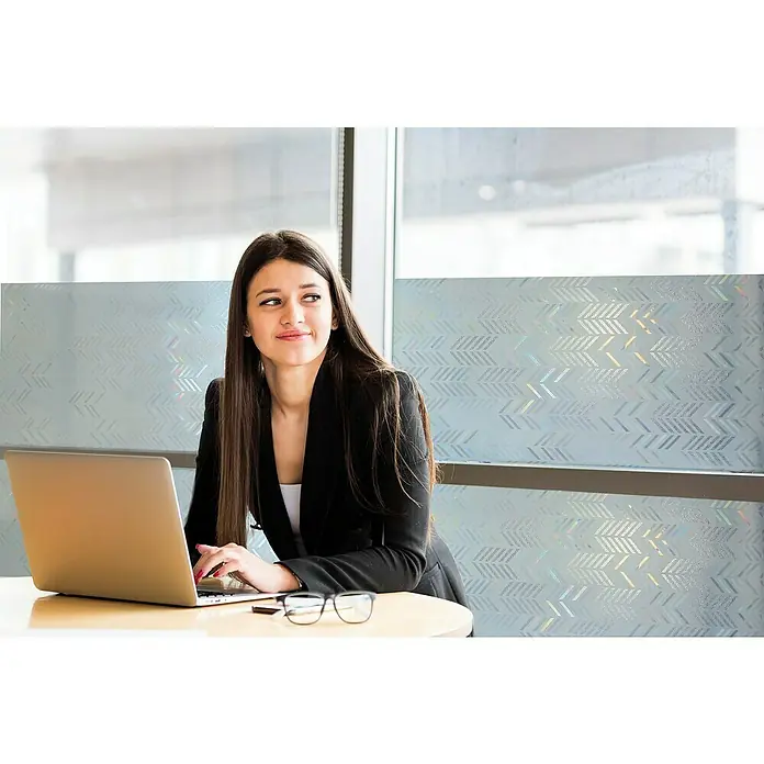 Lámina para ventana al corte Espigas - Transparente - Una joven con blazer está sentada en una mesa con un portátil, sonriendo mientras mira hacia un lado con sus gafas al lado.