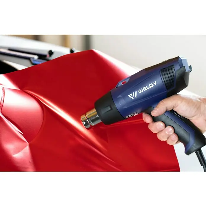 Weldy Heißluftgebläse HG 530-A - 2.300 W, Temperaturbereich: 80 °C - 650 °C - Ein blauer Weldy-Heißluftfön wird verwendet, um eine glänzende rote Folie auf einem Auto anzubringen. Das Werkzeug ist ergonomisch geformt.