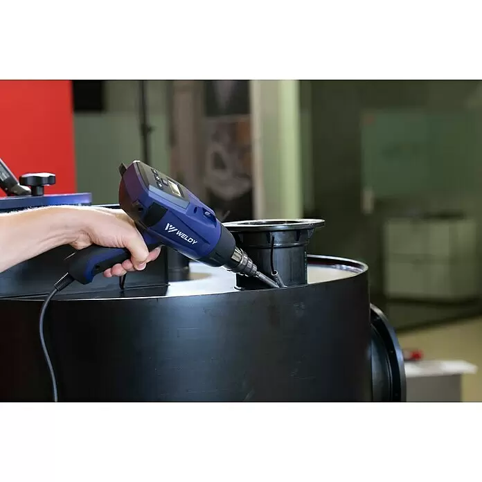 Weldy Heißluftgebläse HG 530-A - 2.300 W, Temperaturbereich: 80 °C - 650 °C - Eine Hand hält ein blau-schwarzes Weldy Heißluftgerät und bearbeitet damit ein großes schwarzes zylindrisches Objekt.