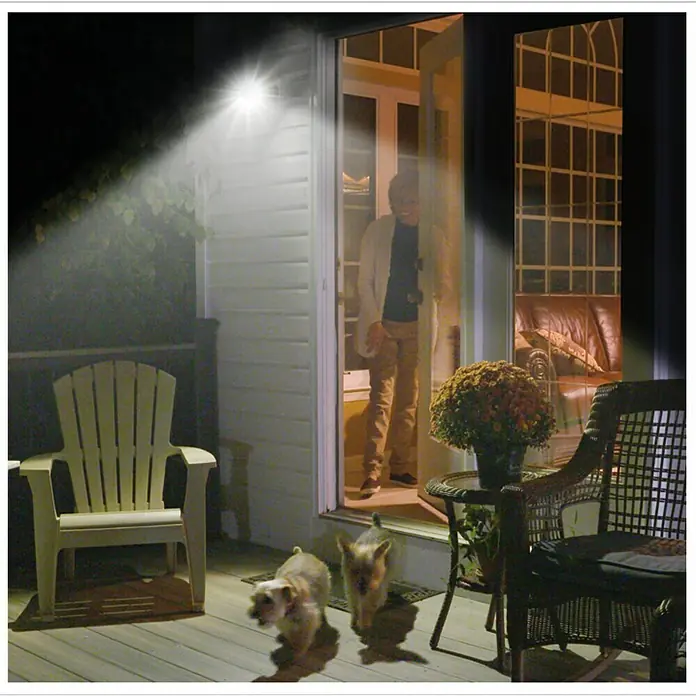 Foco solar Handy Brite - Sensor de luz - Una luz exterior de energía solar ilumina un patio con una silla, una mesa y plantas. Una persona y dos perros están en la puerta.