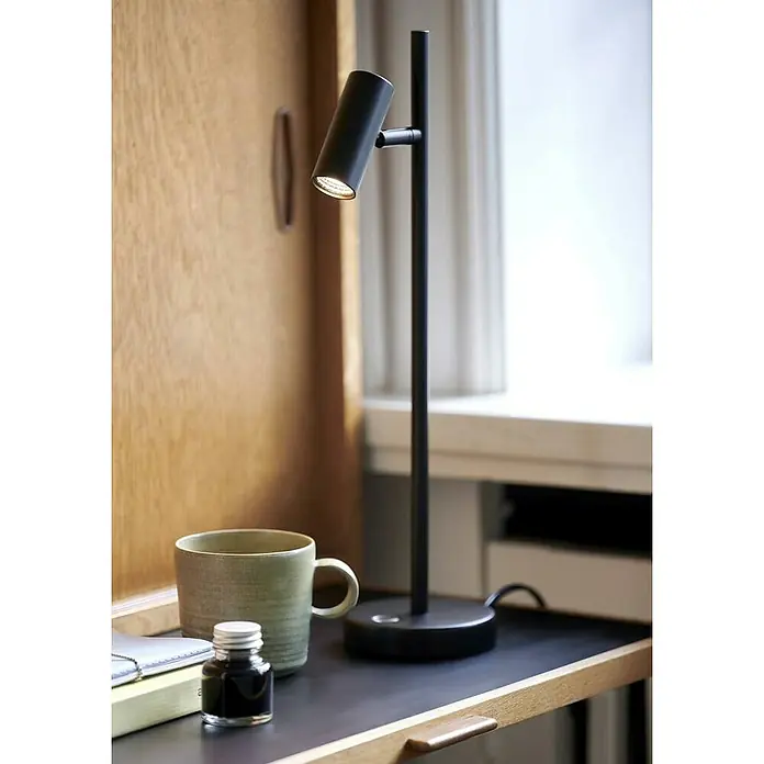 Nordlux Omari Lámpara de sobremesa LED - 3,3 W, L x An x Al: 10 x 15,5 x 40 cm, Negro, Color de luz: Blanco cálido - Una lámpara de escritorio negra y moderna ilumina un escritorio con una taza y un tintero sobre un libro.