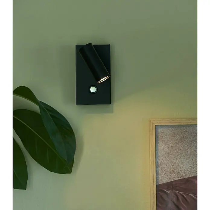 Nordlux Omari Aplique de pared LED - 3,3 W, L x An x Al: 7 x 12 x 12 cm, Negro - Primer plano de una lámpara de pared LED negra con un foco ajustable, que emite una luz cálida, montada en una pared verde.