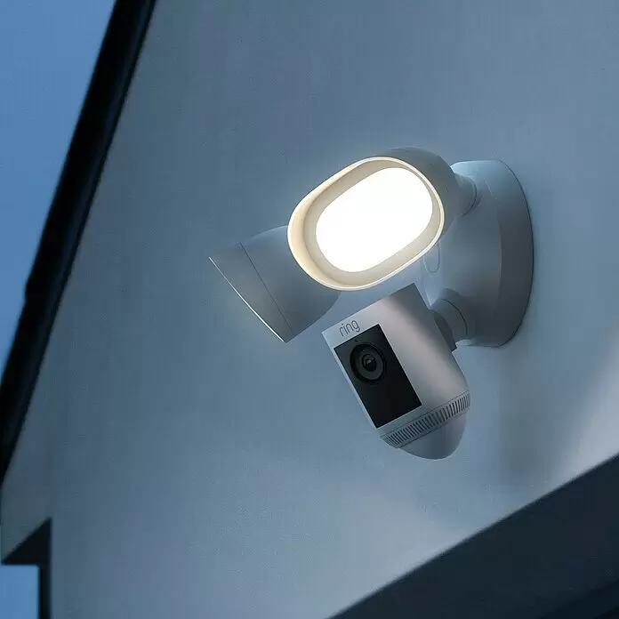 Ring Überwachungskamera Flutlichtkamera Pro Kabel - Weiß, Erfassungswinkel: 140 ° - Weiße Ring Flutlichtkamera mit zwei Lichtern und zentraler Kamera, abends an einem Gebäude zur Sicherheit montiert.