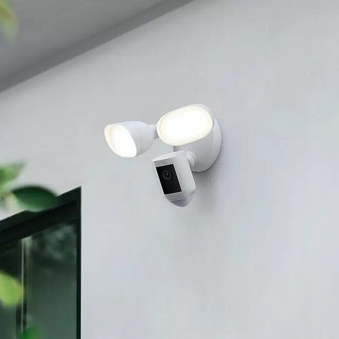 Ring Überwachungskamera Flutlichtkamera Pro Kabel - Weiß, Erfassungswinkel: 140 ° - Weiße Ring Überwachungskamera mit zwei Flutlichtern, an einer Wand montiert.