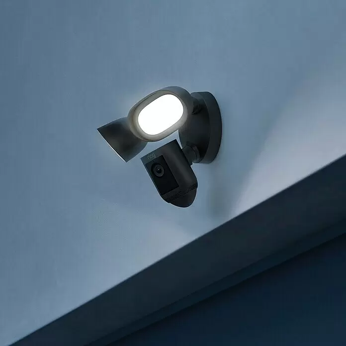 Ring Überwachungskamera Flutlichtkamera Pro Kabel - Schwarz, Netzbetrieben, 2.048 x 1.536 Pixel (2K-HD) - Eine dunkelgraue Ring Floodlight Cam mit zwei hellen Lichtern und einer Kamera, an einer Wand montiert und die Umgebung beleuchtend.