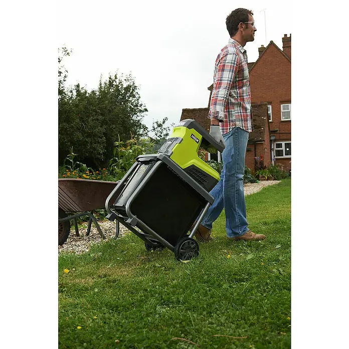 Ryobi Elektro-Leisehäcksler RSH3045U - Max. Aststärke: 4,5 cm, 3.000 W - Ein Mann zieht einen elektrischen Ryobi-Gartenhäcksler 3000W durch einen grünen Garten mit einem Backsteinhaus im Hintergrund.