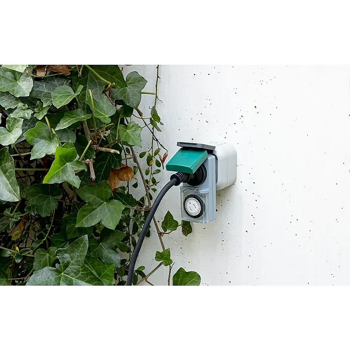 Temporizador para exterior - Mecánico, Gris / Verde, 3.500 W - Primer plano de un temporizador exterior en una pared rodeado de hiedra, destacando las características del producto.