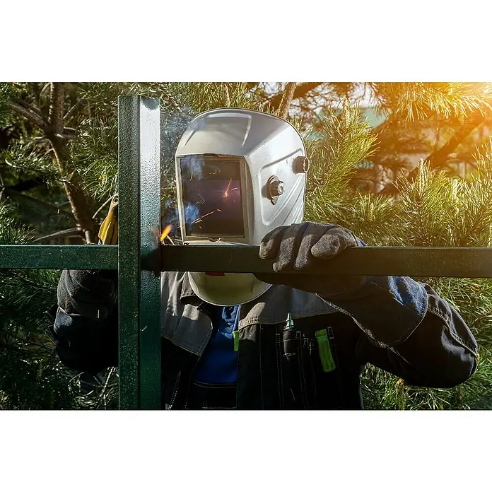 Gys Elektrodenschweißgerät GYSMI 130P - Schweißstrom: 10 A - 130 A, 230 V - Ein Schweißer mit Schutzhelm und Handschuhen, der Metallprofile mit einem Schweißgerät verbindet, im Hintergrund grünes Blattwerk.