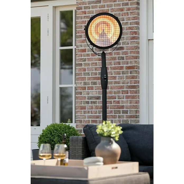 Sunred Halogen-Heizstrahler Indus - Höhe: 210 cm, 2.100 W, Schwarz - Nahaufnahme einer Terrassenheizung mit Standfuß, die Wärme abstrahlt, vor einer Ziegelwand und einer Sitzecke im Freien.