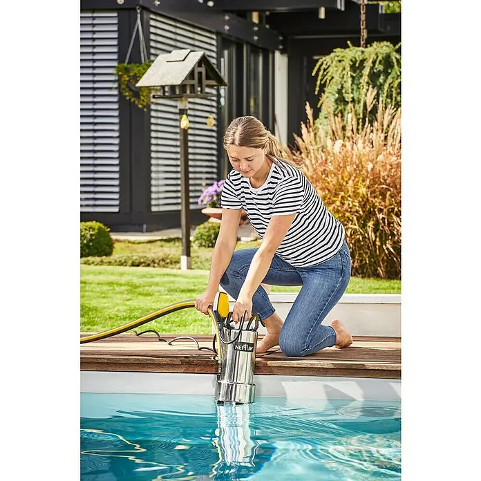Neptun Bomba para aguas residuales GC-DP 9035 N - 950 W, Capacidad de bombeo máx.: 18.200 l/h, Profundidad de inmersión: 5 m - Mujer operando una bomba sumergible Neptun junto a la piscina. La bomba es de acero inoxidable con un mango amarillo, diseñada para bombear agua.