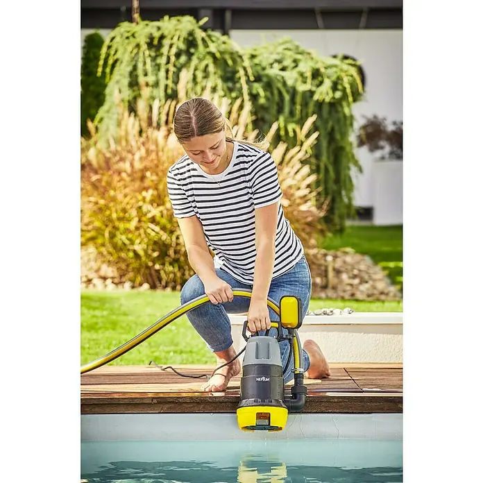 Neptun Bomba para aguas residuales NSP-E 75 - 750 W, Capacidad de bombeo máx.: 15.500 l/h, Profundidad de inmersión: 7 m - Mujer usando una bomba sumergible Neptun para vaciar una piscina en el jardín. La bomba es gris y amarilla y tiene una manguera amarilla.