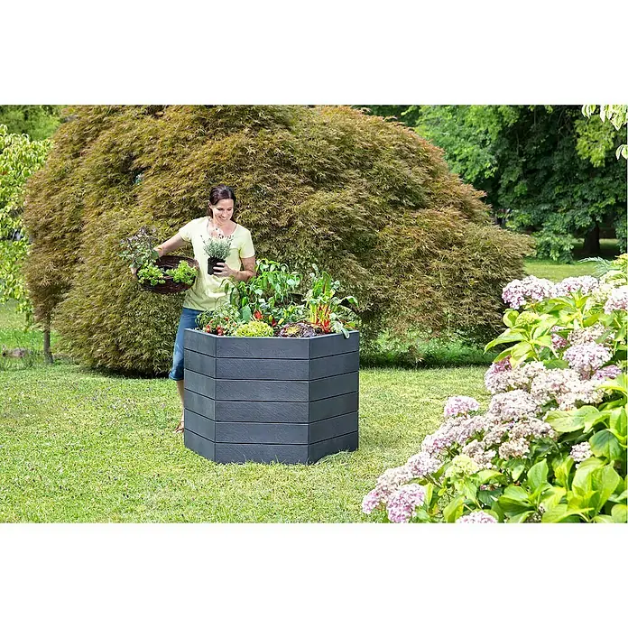 Garantia Hochbeet Ergo L Stone - Ø x H: 110 x 75 cm, Kunststoff, Anthrazit - Eine Frau bepflanzt ein Hochbeet im Garten. Das graue Beet ist mehrschichtig und mit Gemüse bepflanzt, umgeben von Grün.