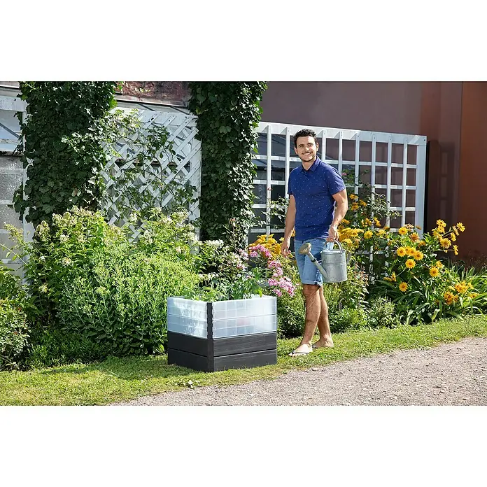 Garantia Hochbeet Ergo Quadro Turbo S Wood - Set - 60 x 60 x 50 cm, Kunststoff, Anthrazit/Braun - Ein Mann mit Gießkanne steht neben einem Hochbeet mit grauem Rahmen und durchsichtigen Pflanzgefäßen, umgeben von üppigem Grün.