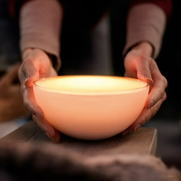 Philips Hue LED-Tischleuchte Go - 6 W, L x B x H: 15 x 15 x 7,9 cm, RGBW - Beleuchtete Schale: Eine Person hält eine leuchtende, runde Schale in ihren Händen, die ein warmes, orangefarbenes Licht ausstrahlt.