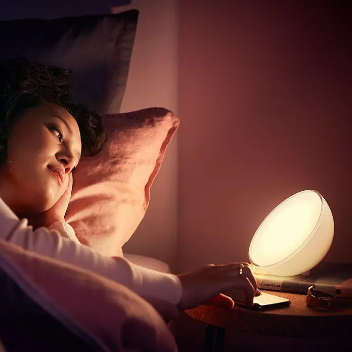 Philips Hue LED-Tischleuchte Go - 6 W, L x B x H: 15 x 15 x 7,9 cm, RGBW - Eine junge Frau liegt im Bett mit einer Philips Wake-up Light Lampe auf ihrem Nachttisch, die ein sanftes Licht spendet.