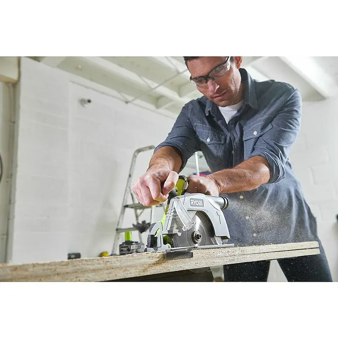 Ryobi ONE+ Akku-Handkreissäge R18CS-0 - 18 V, Ohne Akku, Leerlaufdrehzahl: 4.700 U/min - Ein Mann sägt ein Brett mit einer Ryobi-Kreissäge. Staubpartikel sind in der Luft sichtbar. Er trägt eine Schutzbrille.