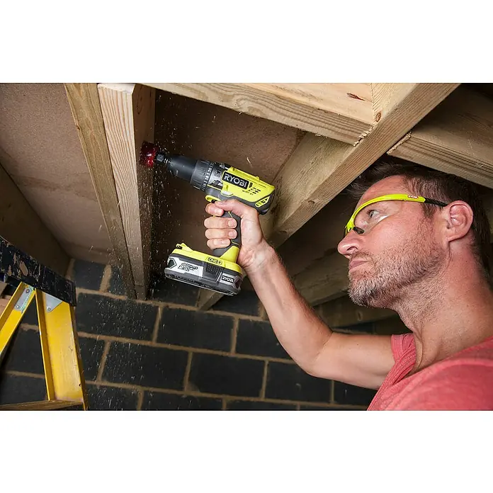 Ryobi ONE+ Taladro atornillador percutor de batería R18PD-0 - 18 V, Sin batería, 50 Nm - Un hombre usa un taladro amarillo RYOBI para perforar un agujero en un techo de madera, las virutas de madera vuelan mientras usa gafas protectoras.