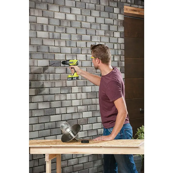 Ryobi ONE+ Taladro atornillador percutor de batería R18PD-0 - 18 V, Sin batería, 50 Nm - Un hombre usa un taladro inalámbrico Ryobi amarillo para perforar un agujero en una pared de ladrillos para instalar una lámpara exterior.