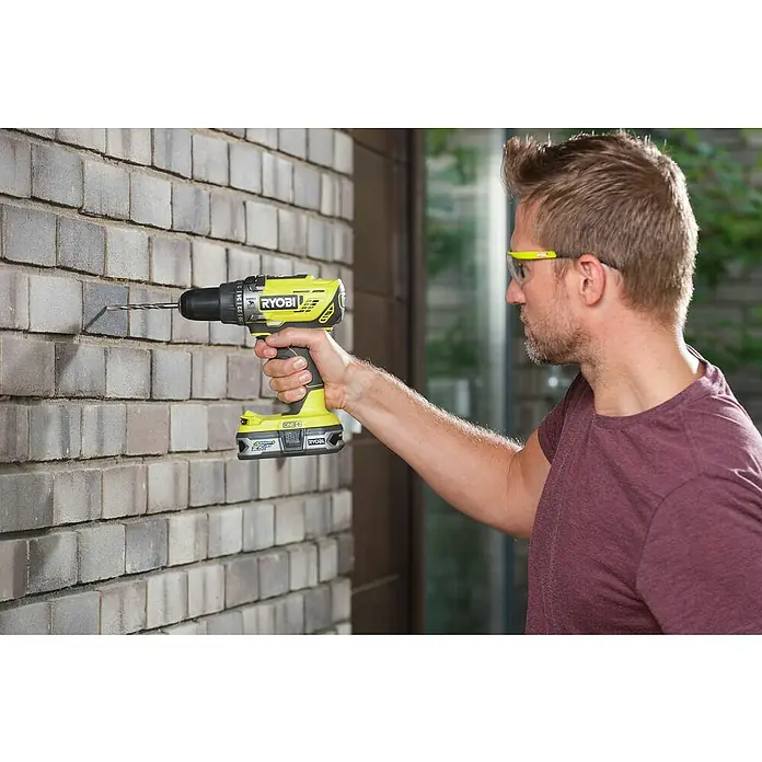 Ryobi ONE+ Taladro atornillador percutor de batería R18PD-0 - 18 V, Sin batería, 50 Nm - Un hombre perfora una pared de ladrillos con un taladro inalámbrico Ryobi amarillo.
