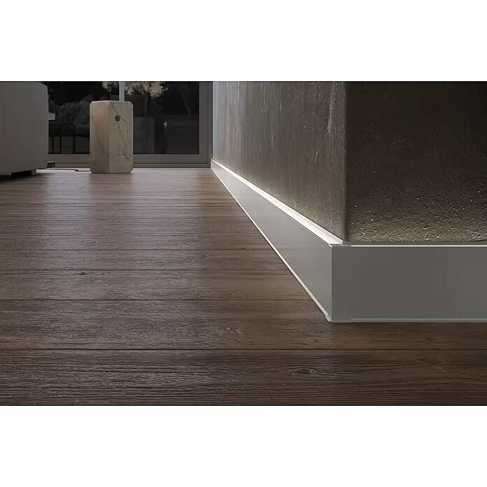 Deckprofil Cubica LS80 Light Up - Aluminium, 2,5 m x 19 mm x 80 mm - Graue Fußleiste mit integrierter LED-Beleuchtung auf Holzboden, beleuchtet eine strukturierte Wand.