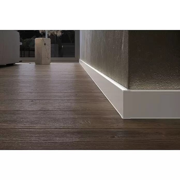 Deckprofil Cubica LS80 Light Up - Weiß, 2,5 m x 19 mm x 80 mm - Weiße Sockelleiste mit integrierter LED-Beleuchtung, die eine strukturierte Wand über dunklem Holzboden beleuchtet.