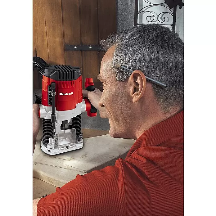Einhell Fresadora eléctrica TC-RO 1155 E - 1.100 W, 11.000 r.p.m. - 30.000 r.p.m., Diámetro del portaherramientas: 6 mm - 8 mm - Un hombre usa una fresadora de inmersión Einhell roja y negra para trabajar en una tabla de madera.