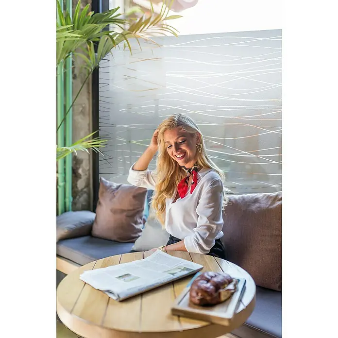 Lámina para ventana Estático Gofrado Mini Rollo Garbi - Transparente - Una mujer rubia está sentada en una mesa de un café, con un periódico y un pastel. Sonríe y lleva un collar rojo.