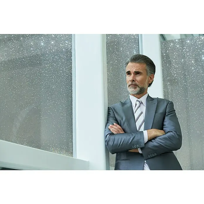 Lámina para ventana Estático Gofrado Mini Rollo Rice - Transparente - Un hombre elegante con un traje gris apoyado pensativamente contra una ventana. Lleva una camisa a rayas y una corbata a juego.