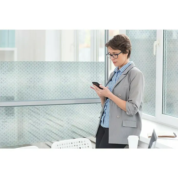 Lámina para ventana al corte estático gofrado fix Rissani - Transparente - Una mujer con blazer escribe en su teléfono inteligente mientras se apoya en una ventana, ambiente de oficina moderna.