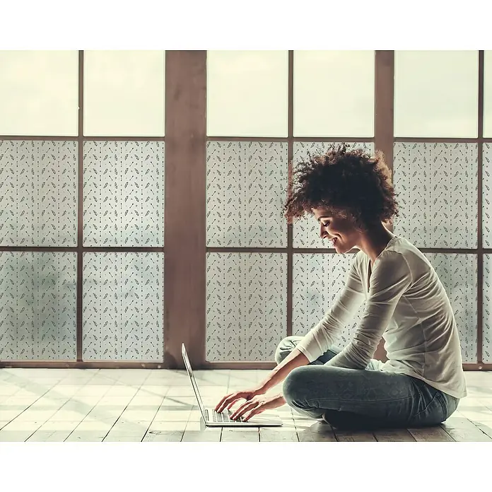 Lámina para ventana al corte digital fix Feather - Blanco/Negro - Una mujer sentada en el suelo usa una laptop frente a una lámina para ventanas con un sutil patrón de flechas para privacidad.