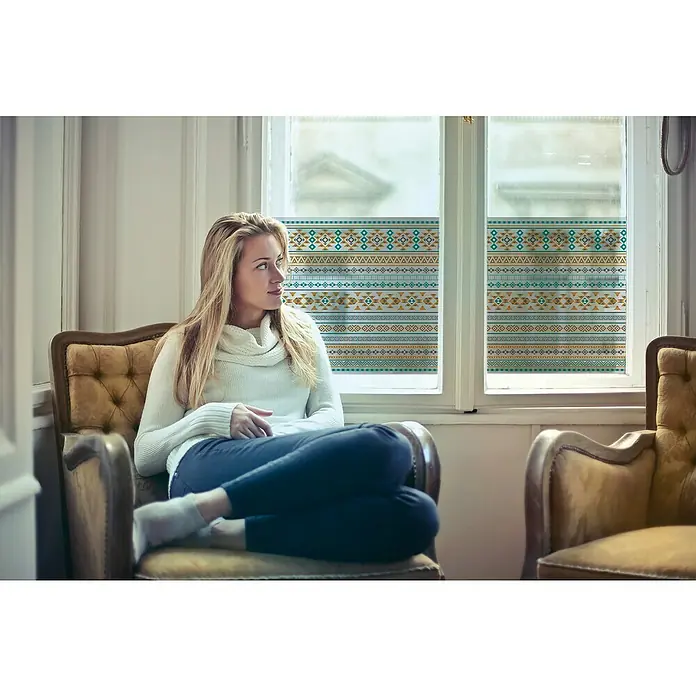 Lámina para ventana al corte fix maya Teal - Multicolor - Una mujer rubia se sienta relajada en un sillón junto a una ventana con una película decorativa, que añade privacidad y estilo.