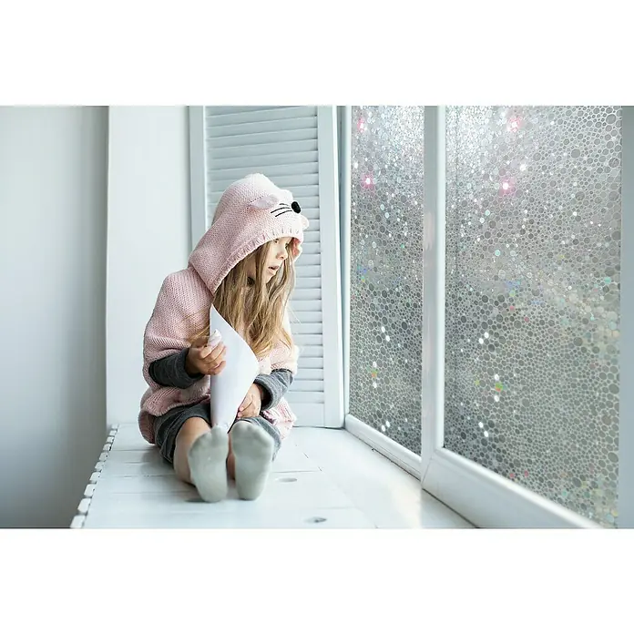 Lámina para ventana al corte gofrado multilámina fix Codol - Transparente - Una niña se sienta en la ventana con una película decorativa que dispersa la luz y aporta privacidad. La película tiene un interesante patrón de burbujas.